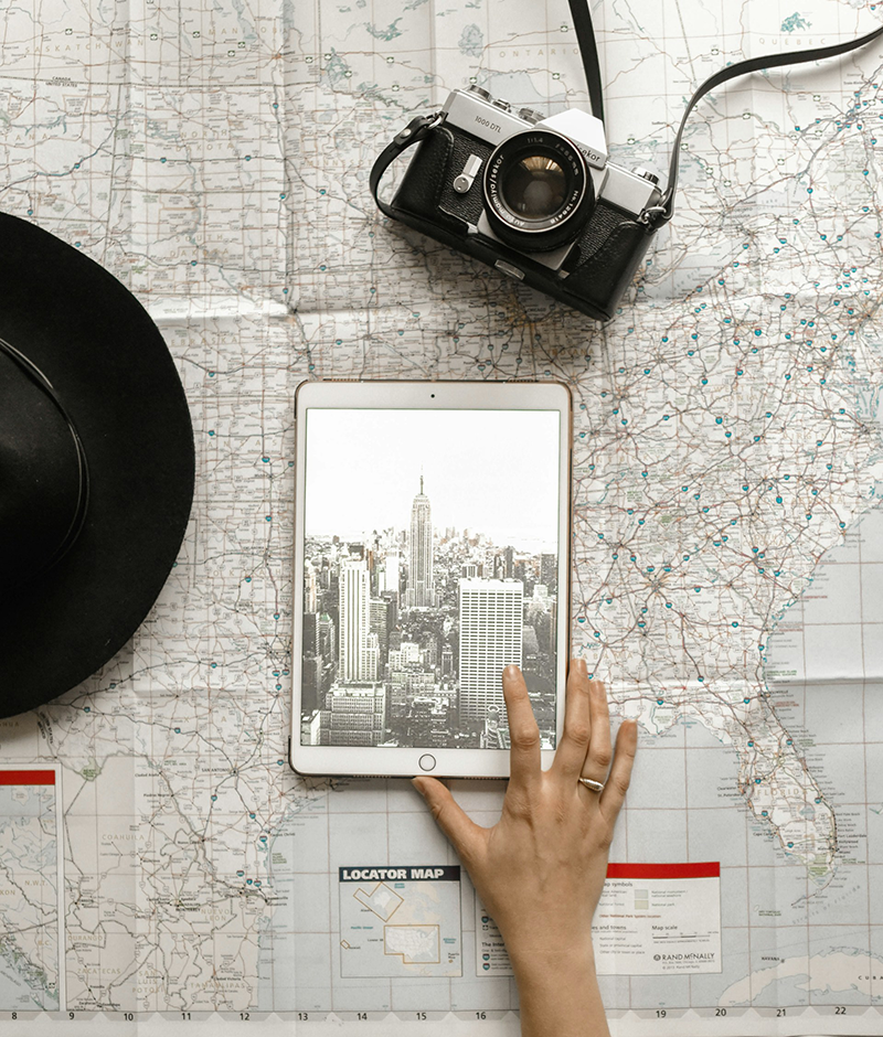 Hand on a tablet showing a city skyline, resting on a road map next to a camera and a hat